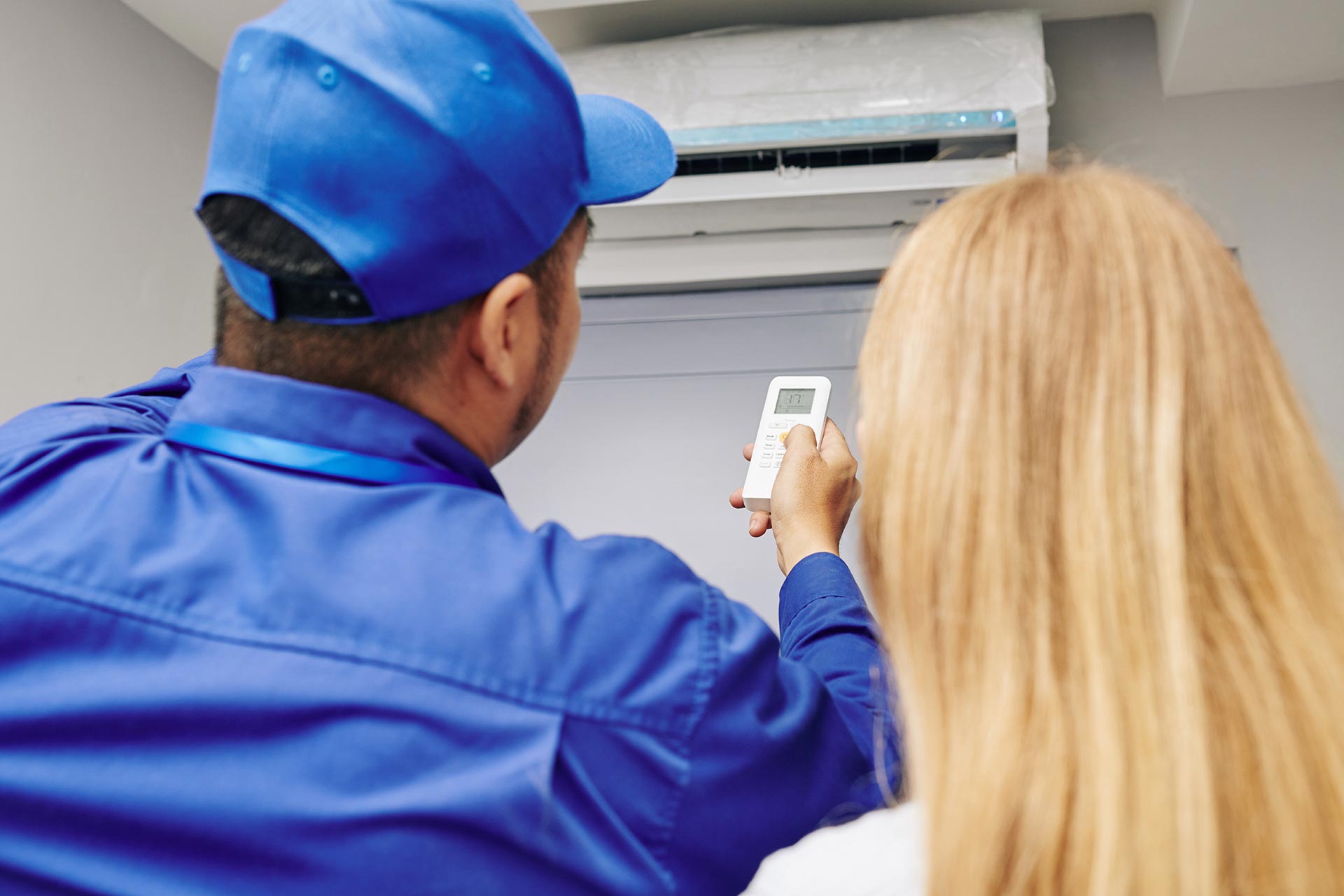 A technician in a blue uniform demonstrates a remote control to a person with blond hair, in front of a wall-mounted air conditioner unit indoors.