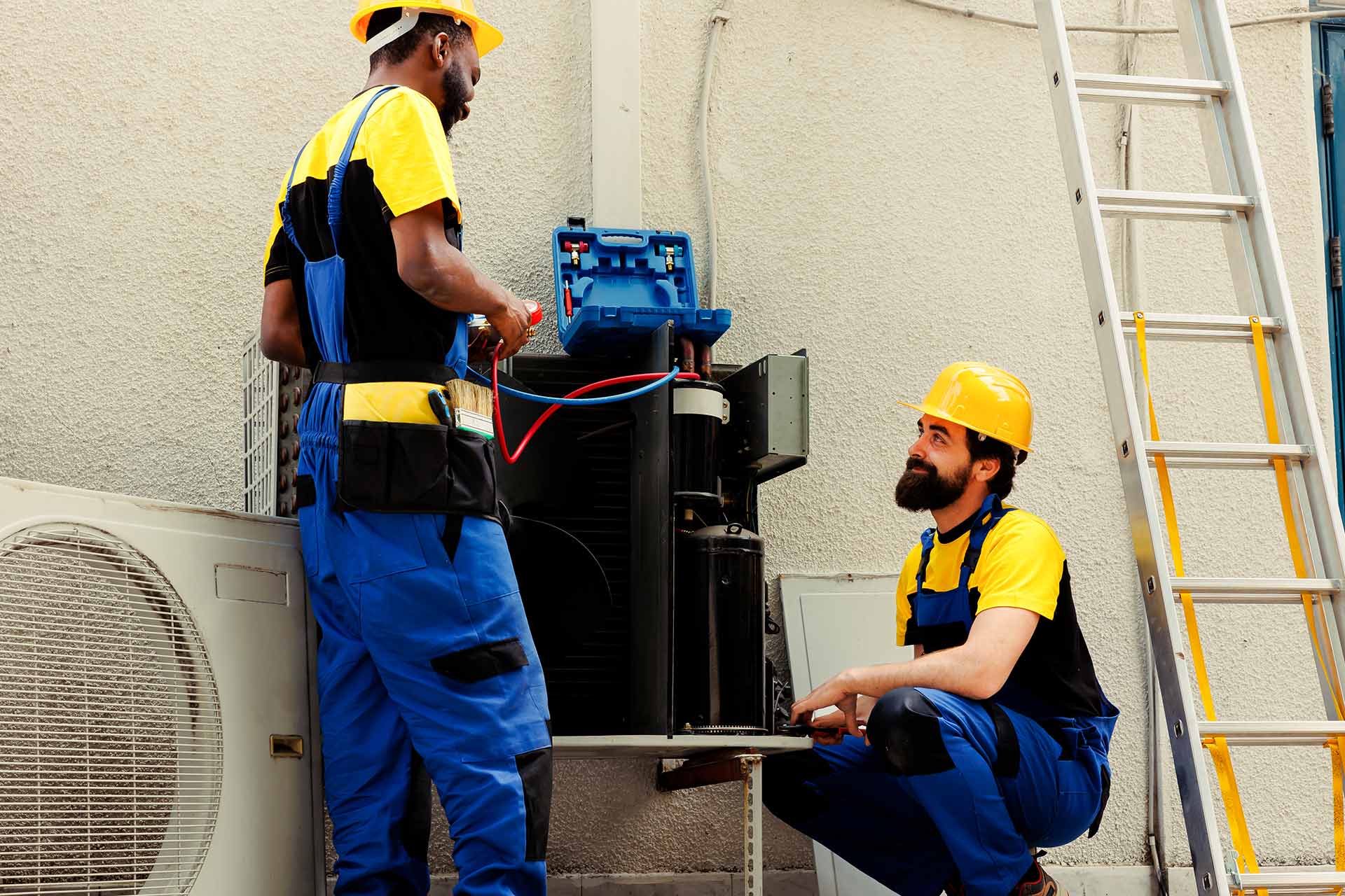 Two workers in yellow helmets and blue uniforms repair an outdoor air conditioning unit. One stands holding tools while the other crouches, smiling. A ladder is nearby.