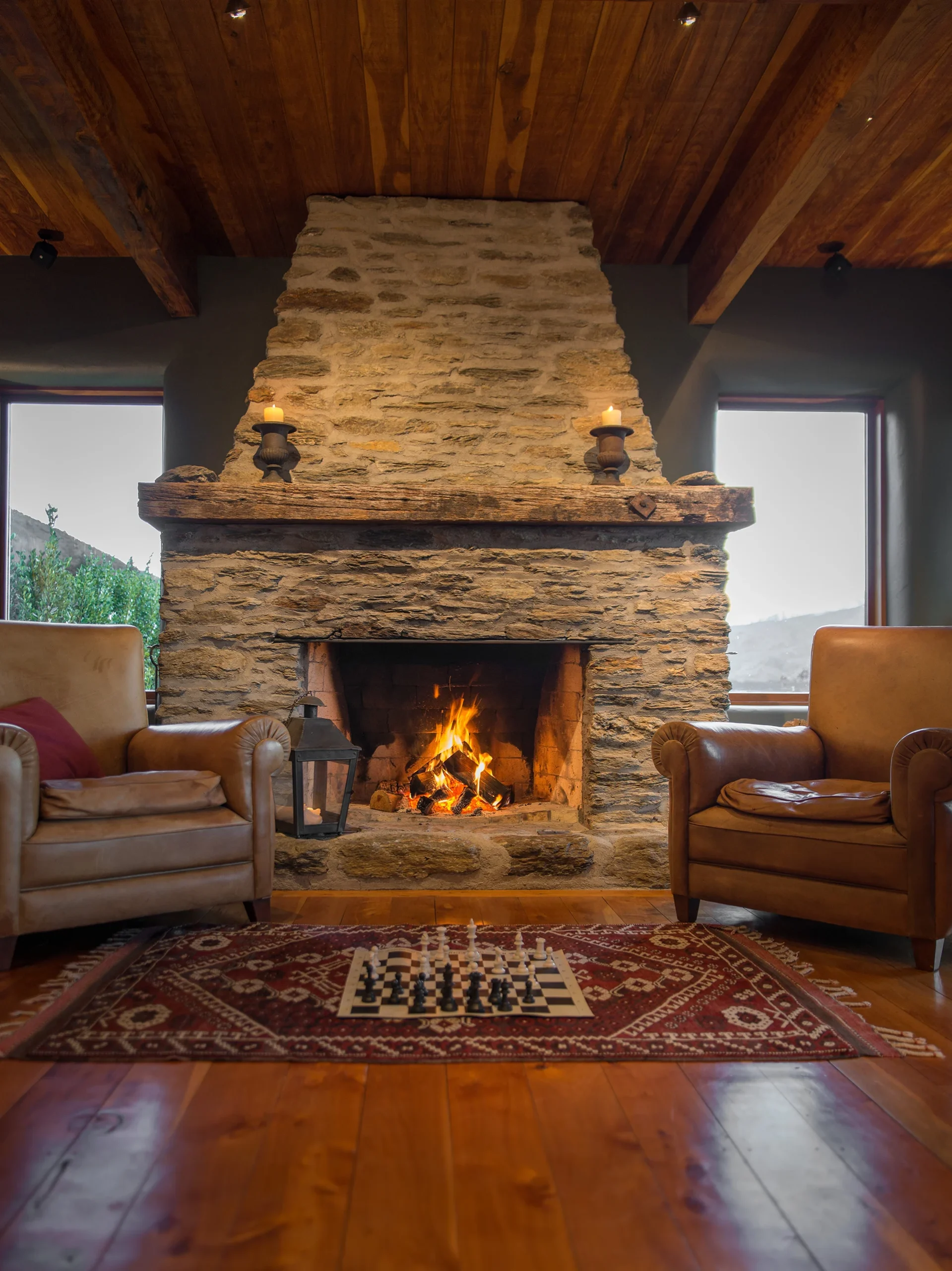 A cozy living room featuring a large stone fireplace, two leather armchairs, a wooden ceiling, and a chess set on a patterned rug.
