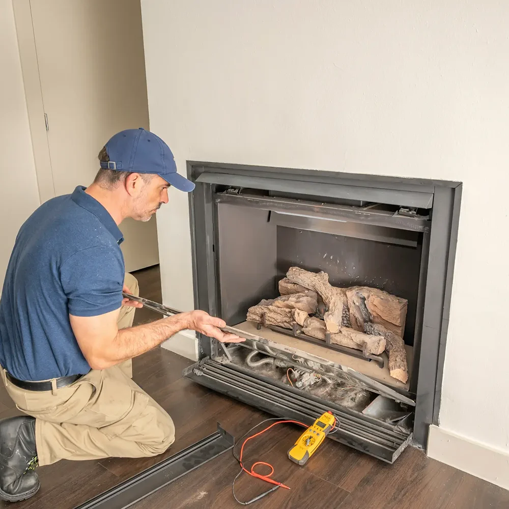 A technician kneeling on a dark wood floor while inspecting and repairing a modern built-in gas fireplace.