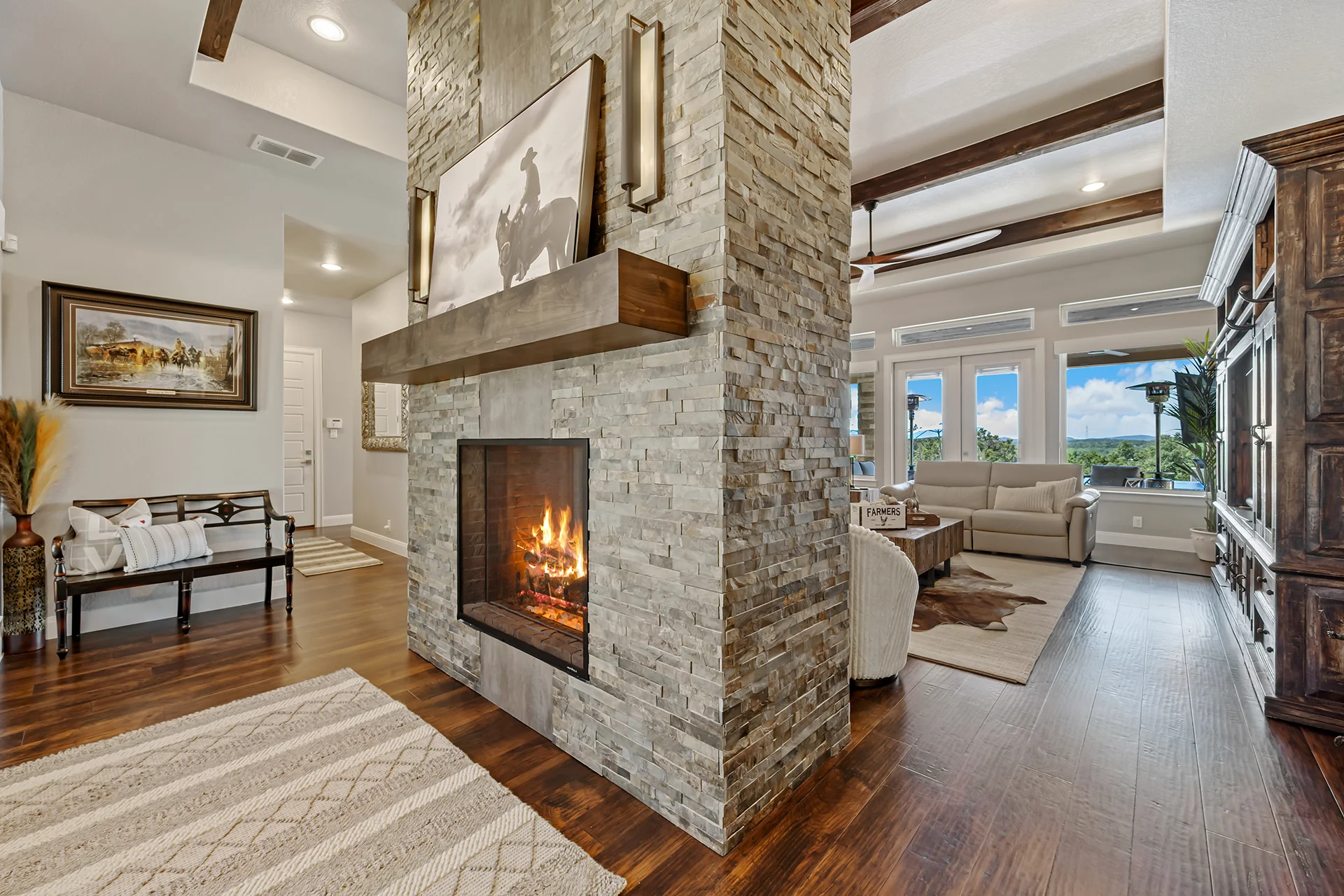 A spacious living room featuring a double-sided stone fireplace with a wooden mantel, dark hardwood floors, vaulted ceilings with wooden beams, and a view of a seating area with a sofa.